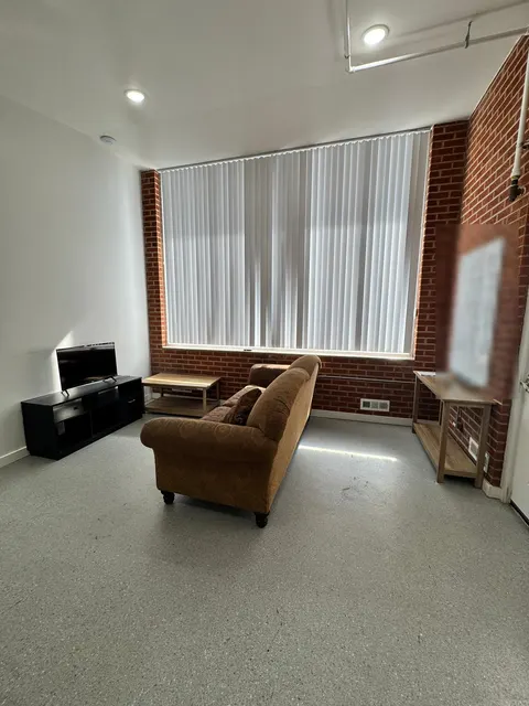 Common room at the 676 Shoup Ave W house with a sofa, TV stand and tall window with blinds between brick walls