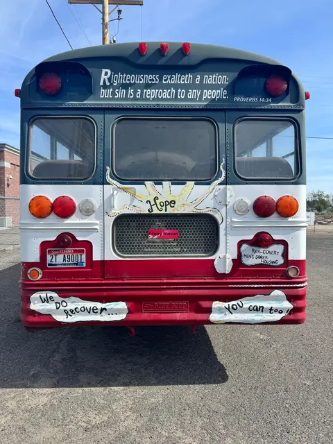 Rear of the painted Realcovery bus with hand-painted bumper signs, parked on gravel at the 676 Shoup Ave W house